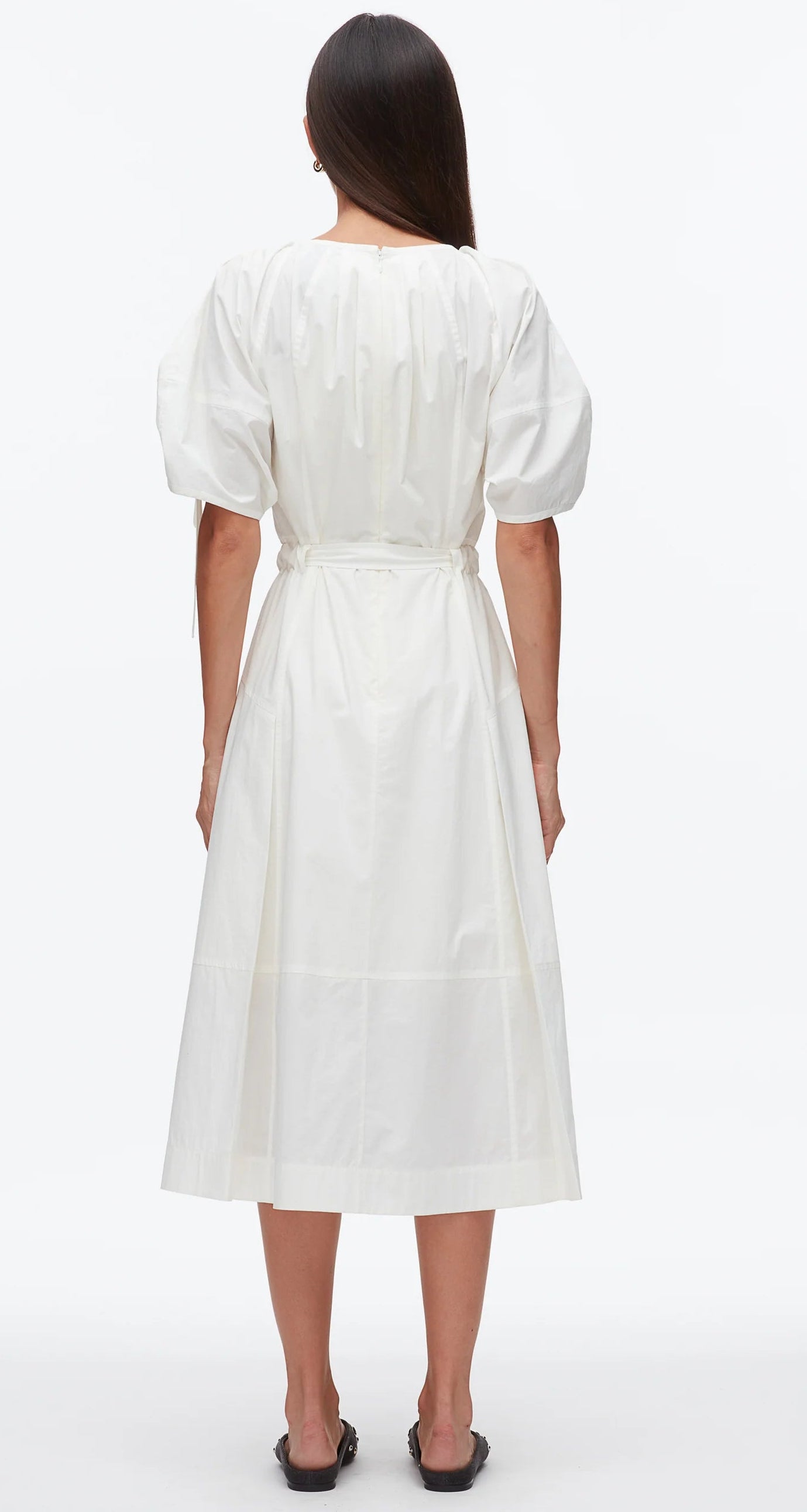 A woman faces away, wearing the 3.1 Phillip Lim Ruched Sleeve Midi Dress in white cotton poplin with puffed short sleeves and a D-ring belt at the waist, paired with black sandals.