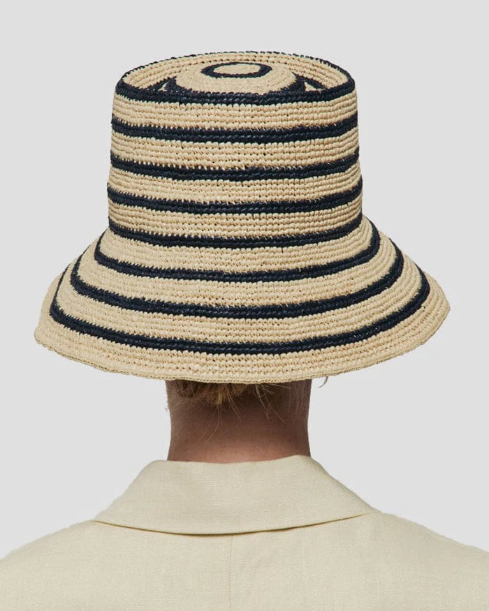 A person is seen from behind wearing the Janessa Leone Felix Harbor Hat, a beige and black striped hand-crocheted raffia bucket hat, paired with a light-colored collared garment.