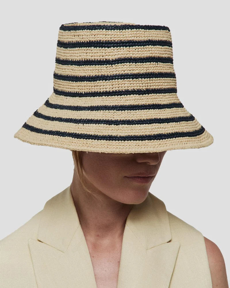 A person wearing a cream blazer and the Janessa Leone Felix Harbor Hat, a wide-brimmed hand-crocheted raffia bucket hat that covers their eyes, against a plain background.
