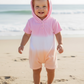 A toddler wearing the Feather 4 Arrow Baby Girls’ Finn Romper stands barefoot on the beach, smiling, with the ocean waves in the background.