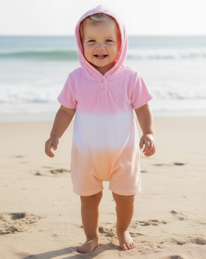A toddler wearing the Feather 4 Arrow Baby Girls’ Finn Romper stands barefoot on the beach, smiling, with the ocean waves in the background.