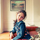 A young girl with long brown hair sits on a leather couch, wearing the Louis Louise Girls' Sybillou Light Denim Tunic with orange dots and a Peter Pan collar, paired with red pants. A framed picture and metallic lamp are in the background.