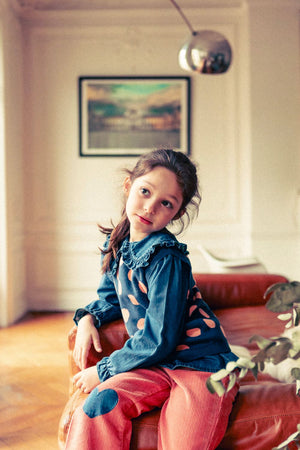 A young girl with long brown hair sits on a leather couch, wearing the Louis Louise Girls' Sybillou Light Denim Tunic with orange dots and a Peter Pan collar, paired with red pants. A framed picture and metallic lamp are in the background.