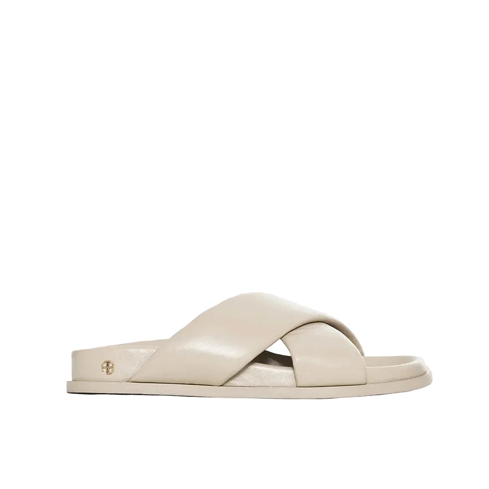 The Emi Slides by Anine Bing are beige flat sandals with padded crisscross straps and a flat sole, finished with a small metallic detail near the heel.
