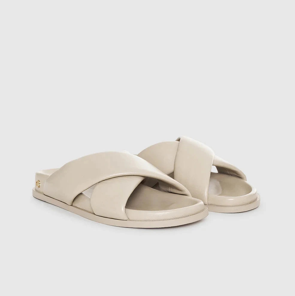 The Emi Slides by Anine Bing are beige flat sandals featuring padded cross-over straps and flat soles, shown on a plain light background.
