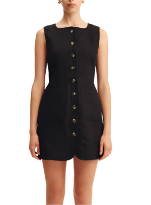 A woman wearing the POSSE Emma Mini Dress—a sleeveless black linen dress with a high neckline, front buttons, and two large front pockets—stands against a white background.