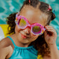 A young girl in a blue UV protection swimsuit sits by the pool, smiling as she holds her Bling2O Kids' Enchanted Shoreline Shimmer Seashell Goggles.