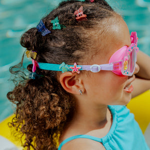 A young child with curly hair, colorful hair clips, and Bling2O Kids' Enchanted Shoreline Shimmer Seashell Goggles sits on a yellow flotation device in a pool, showcasing playful swimwear perfect for summer fun.