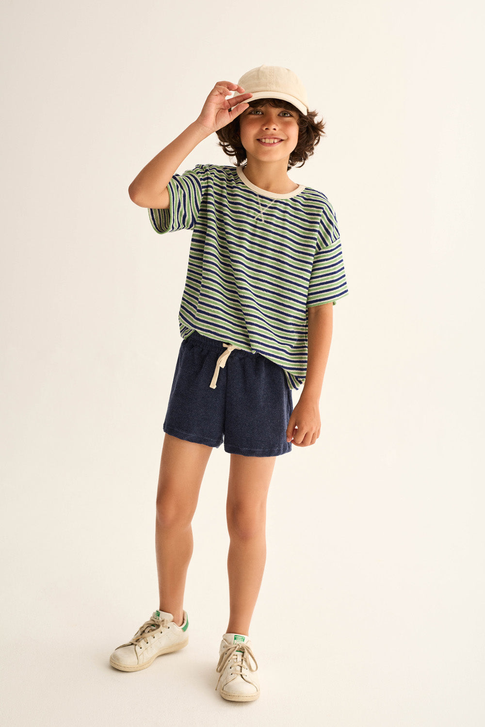 A child wears the Cozmo Boys' Enea Terry T-Shirt with navy shorts, white sneakers, and a beige cap, standing against a plain light background while smiling and holding the cap's brim.
