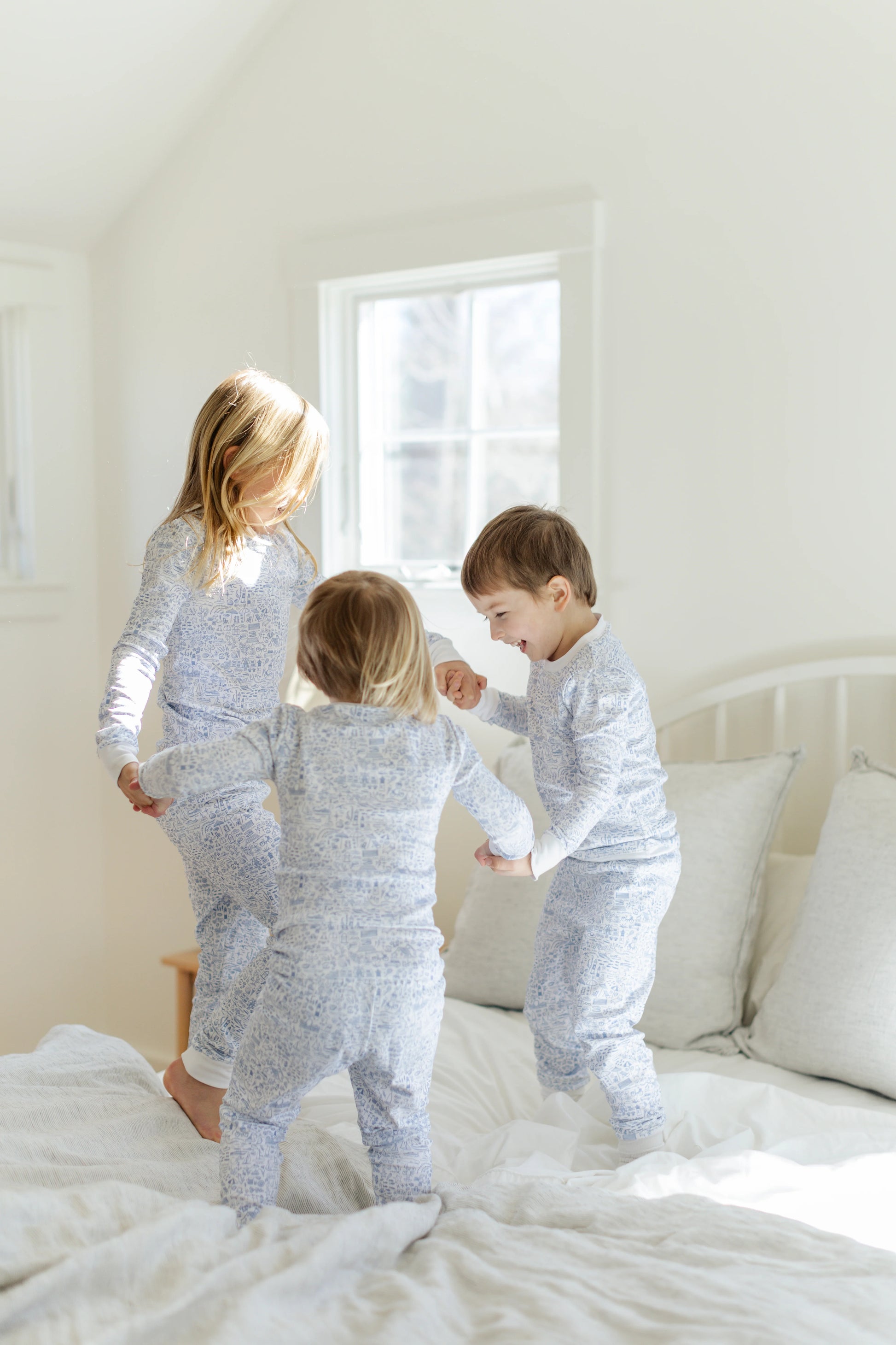 Three young children in matching Joy Street Baby Seaside Two Piece Pajamas play together on a sunlit bed, making memories and celebrating summer traditions.
