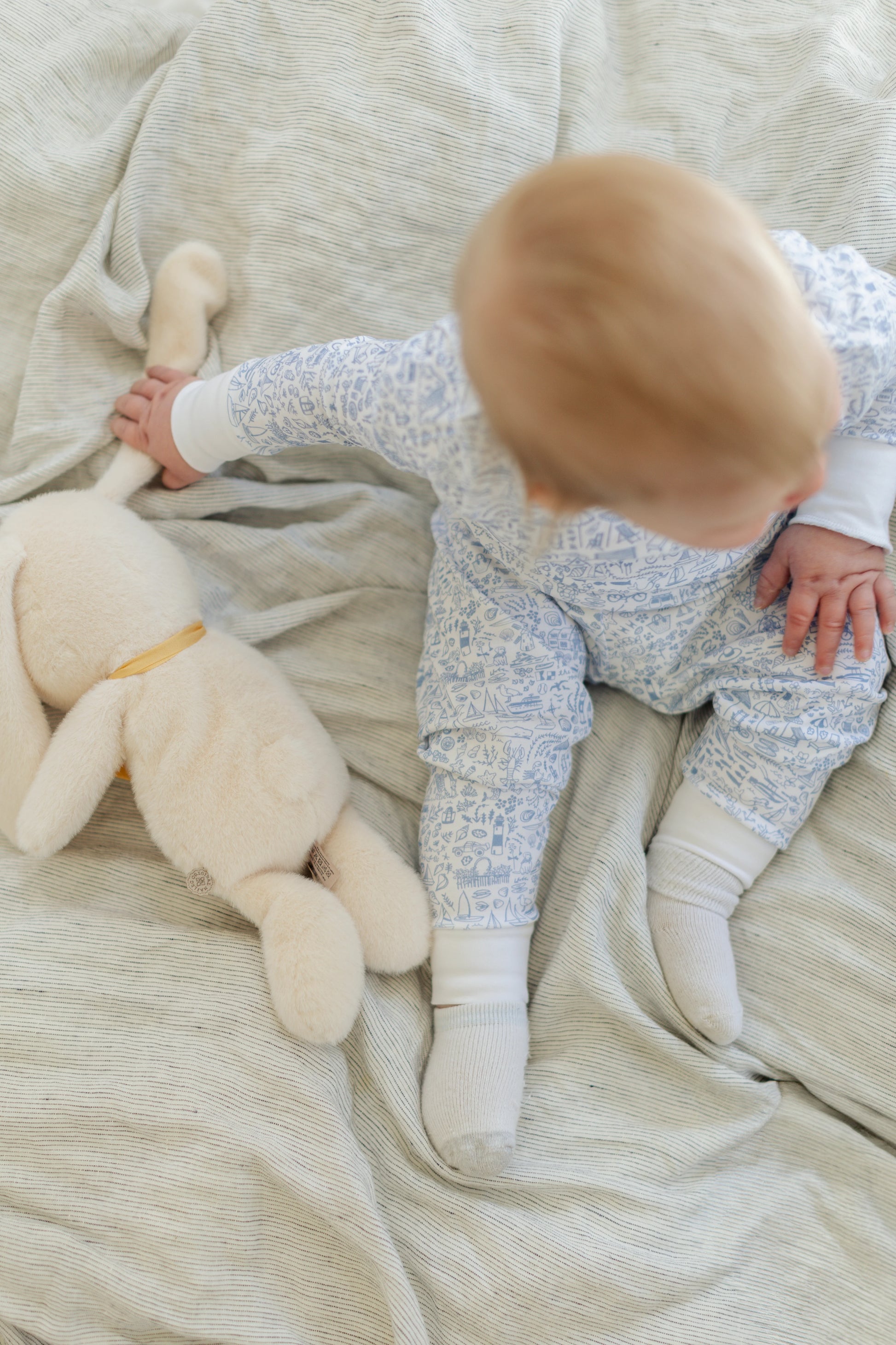 A baby in the Joy Street Baby Seaside Zip Onesie sits on a blanket beside a plush toy, reaching for its tail—capturing that sunny seaside vibe.