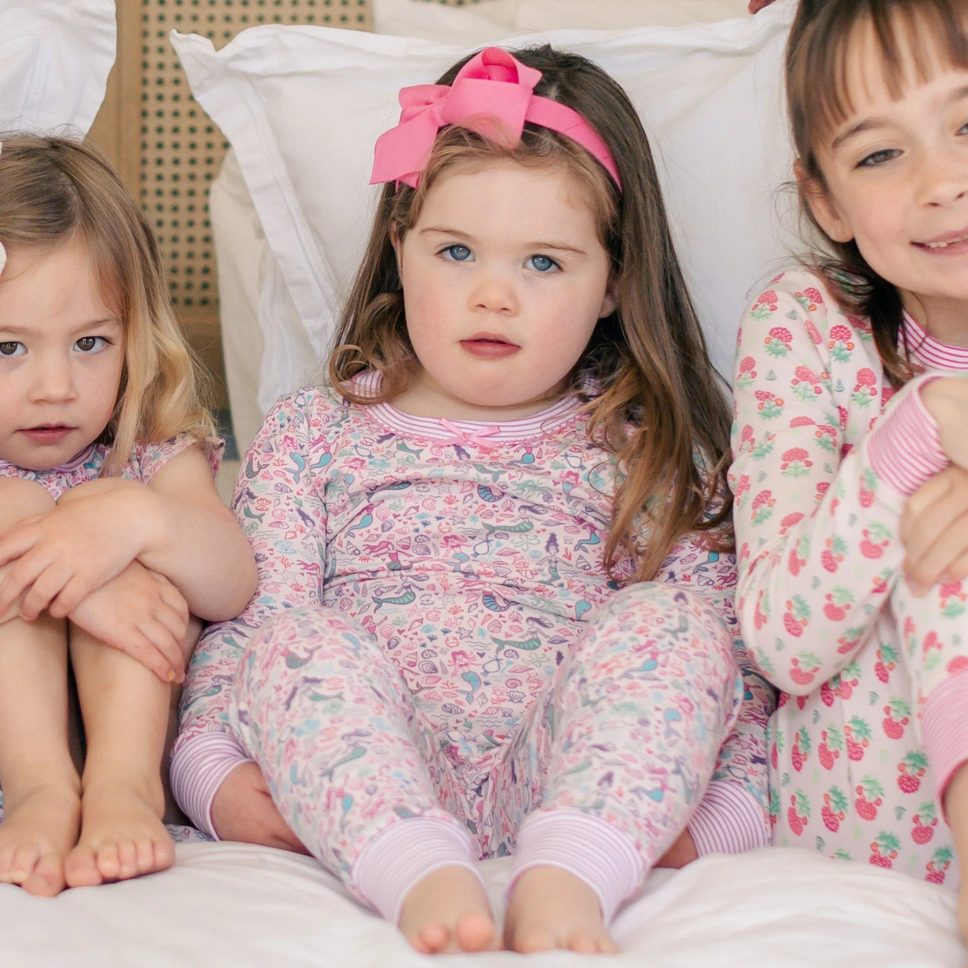 Three young girls in patterned Joy Street pajamas sit together on a bed; the middle girl wears a pink headband, and one set is the Girls' Mermaids Two Piece Pajamas featuring hand-drawn sea creatures.