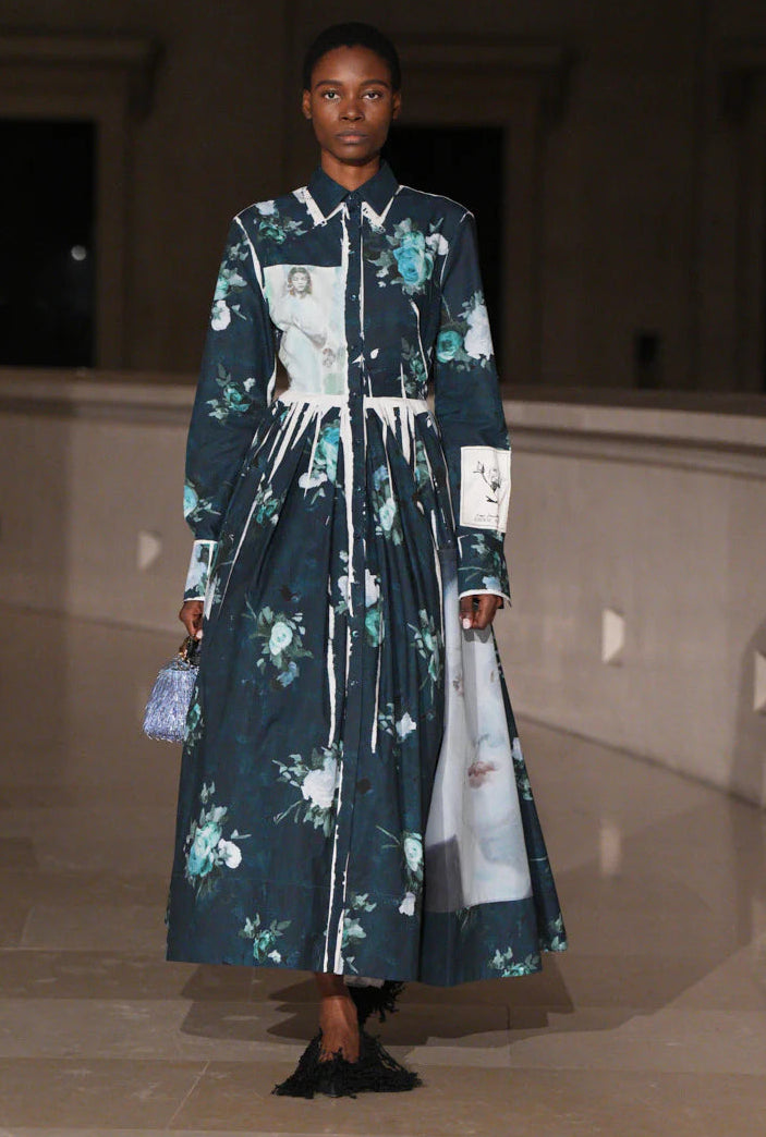 A model walks an indoor runway in the Erdem Long Sleeve Abstract Rose Shirt Dress, featuring a collared design with abstract rose print, and carries a small textured handbag.