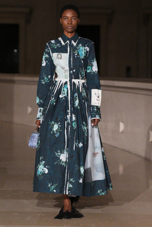 A model walks an indoor runway in the Erdem Long Sleeve Abstract Rose Shirt Dress, featuring a collared design with abstract rose print, and carries a small textured handbag.