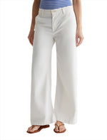 A person wearing white AG Jeans Caden Wide Leg trousers made from Stretch Cotton Tencel Twill, a blue and white striped top, and brown sandals stands against a white background.