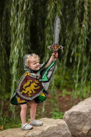 A young child, dressed as a knight, stands on a rock outdoors with a willow tree in the background, smiling and holding the Great Pretenders Knights Honor Foam Sword aloft—fully immersed in imaginative play.
