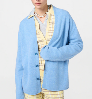 A person wearing the Guest in Residence Everywear Cardigan in light blue with a shawl collar, layered over a yellow plaid shirt and matching pants, accessorized with a beaded necklace.