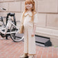 A young girl with long red hair stands on a brick sidewalk holding a book. She wears Rylee & Cru Girls' Pointelle Knit Pants with an elastic waistband and crochet lace hem. A white bicycle is visible in the background.