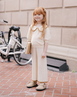 A young girl with long red hair stands on a brick sidewalk holding a book. She wears Rylee & Cru Girls' Pointelle Knit Pants with an elastic waistband and crochet lace hem. A white bicycle is visible in the background.