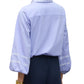 A person shown from the back wearing the Figue Elaine Top, a light blue cotton poplin blouse with blouson sleeves and embroidered collar and cuffs, paired with dark pants and gold bracelets.