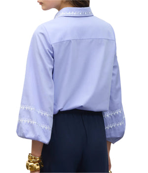 A person shown from the back wearing the Figue Elaine Top, a light blue cotton poplin blouse with blouson sleeves and embroidered collar and cuffs, paired with dark pants and gold bracelets.