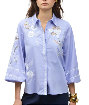 A woman wears the Figue Elaine Top, a light blue cotton poplin blouse with white floral embroidery, an embroidered front placket, three-quarter blouson sleeves, and lace cut-out details.