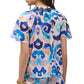 A woman is shown from the back wearing the Figue Elena Top, a short-sleeve blouse with a colorful abstract blue, pink, and purple pattern and smocked neckline. She accessorizes with gold bracelets on her left wrist.