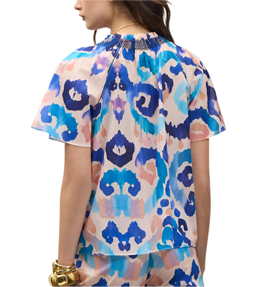 A woman is shown from the back wearing the Figue Elena Top, a short-sleeve blouse with a colorful abstract blue, pink, and purple pattern and smocked neckline. She accessorizes with gold bracelets on her left wrist.