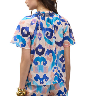 A woman is shown from the back wearing the Figue Elena Top, a short-sleeve blouse with a colorful abstract blue, pink, and purple pattern and smocked neckline. She accessorizes with gold bracelets on her left wrist.