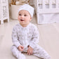 A baby wears the Kissy Kissy Baby Elephant Esprit Printed Zip Footie while sitting on a light nursery floor, surrounded by a crib and stuffed animals.