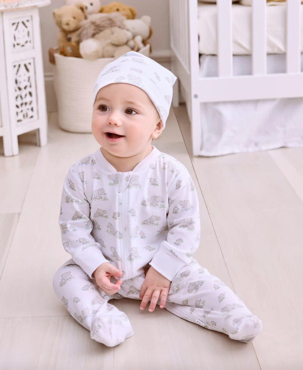 A baby wears the Kissy Kissy Baby Elephant Esprit Printed Zip Footie while sitting on a light nursery floor, surrounded by a crib and stuffed animals.