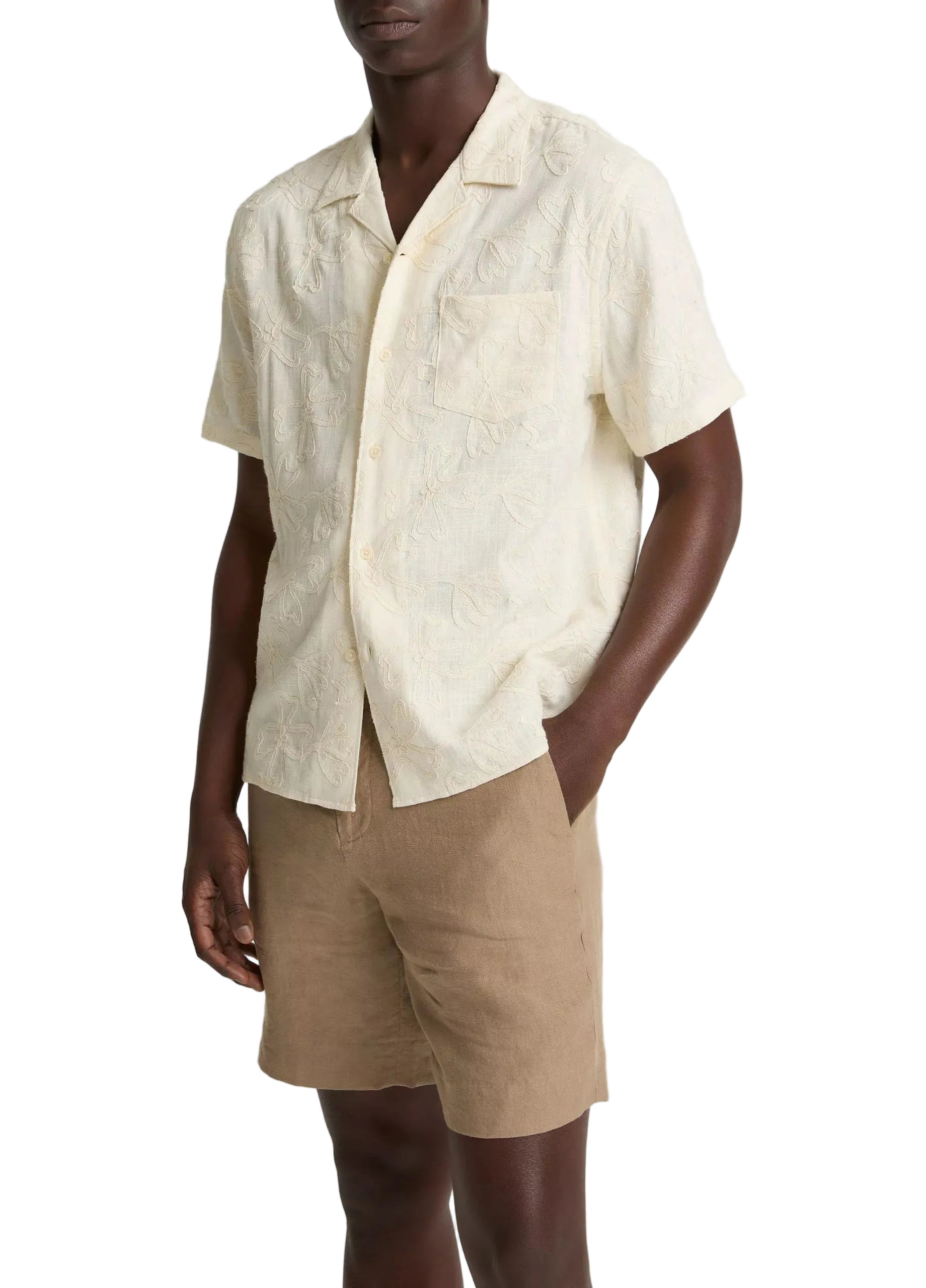 A person wears the vince Embroidered Floral Shortsleeve Cabana Shirt, cream-colored with a subtle floral pattern, paired with tan shorts and standing with one hand in their pocket.