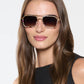 A woman with long brown hair wears Krewe’s Emery Sunglasses, featuring large square unisex frames, and a sleeveless black top, posing against a plain light background.
