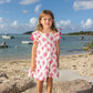 A young girl in the Mer Girls' Sophie Tassel Dress with a tiered skirt stands barefoot on a beachside rock, as boats float nearby under a partly cloudy sky.