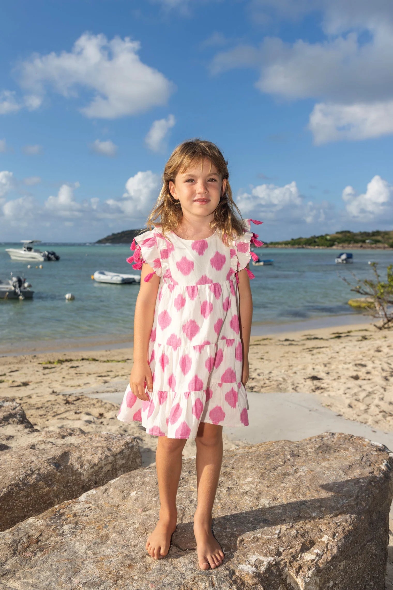 A young girl in the Mer Girls' Sophie Tassel Dress with a tiered skirt stands barefoot on a beachside rock, as boats float nearby under a partly cloudy sky.
