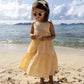 A young child wearing the Early Sunday Girls' Alice Dress with a tiered silhouette and sunglasses stands barefoot on a sandy beach. The cotton dobby fabric flows gently in the breeze, with the ocean and distant islands forming a picturesque backdrop.