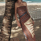 A woman in the Johanna Ortiz Paraiso Agreste Ankle Dress with a strapless neckline leans against a palm tree on a sandy beach, straw bag beside her—ocean and white fence in the background—ideal for summer soirées.