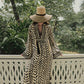 Wearing the Squared Away Tunic Dress by Johanna Ortiz, a person with structured shoulders and a wide-brimmed straw hat sits on a white balcony railing, surrounded by lush green plants and trees in the background.