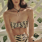 A woman sits outdoors in front of a white lattice, wearing the Johanna Ortiz Sea Sprite & Taita Bikini—a strapless, high-waisted set with a black and beige animal print.