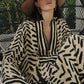 Wearing the Squared Away Tunic Dress by Johanna Ortiz—a patterned beige and black style with structured shoulders—a woman sits on a black wicker chair, wide-brim hat atop her head, surrounded by lush greenery.