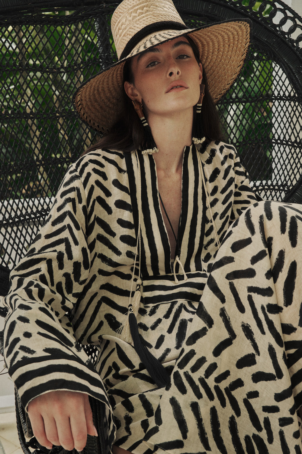 Wearing the Squared Away Tunic Dress by Johanna Ortiz—a patterned beige and black style with structured shoulders—a woman sits on a black wicker chair, wide-brim hat atop her head, surrounded by lush greenery.