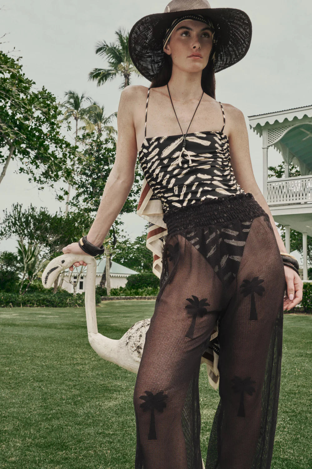 A woman outdoors in a wide-brimmed hat and the Whispering Tides One Piece by Johanna Ortiz leans on a white flamingo statue.