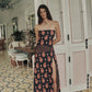 A woman stands barefoot indoors on patterned tile near white paneled doors and a mirror, surrounded by light fixtures and plants. She is wearing the Under The Sun Ankle Skirt by Johanna Ortiz, featuring a bold red and black pattern reminiscent of an abstract JO print.