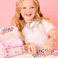 A smiling girl decorates herself and her jewel-covered Super Smalls Mini Vanity Gemify Craft Kit with colorful gem stickers, all set against a pink background.