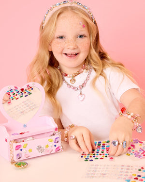 A smiling girl decorates herself and her jewel-covered Super Smalls Mini Vanity Gemify Craft Kit with colorful gem stickers, all set against a pink background.