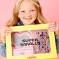 A smiling girl with freckles holds a yellow "Super Smalls" box containing the Mini Vanity Gemify Craft Kit, ready to decorate with colorful gem stickers and create her own sparkling station against a pink background.