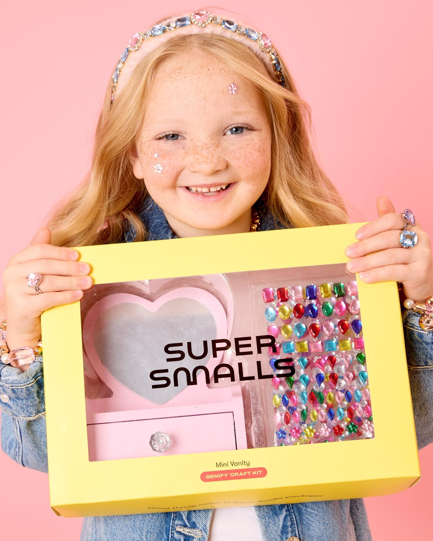A smiling girl with freckles holds a yellow "Super Smalls" box containing the Mini Vanity Gemify Craft Kit, ready to decorate with colorful gem stickers and create her own sparkling station against a pink background.