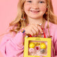 A smiling child with long blonde hair and colorful jewelry holds a yellow Super Smalls gift box containing lip gloss, rings, and the Paint it Heartfelt Mini Nail Kit against a pink background.