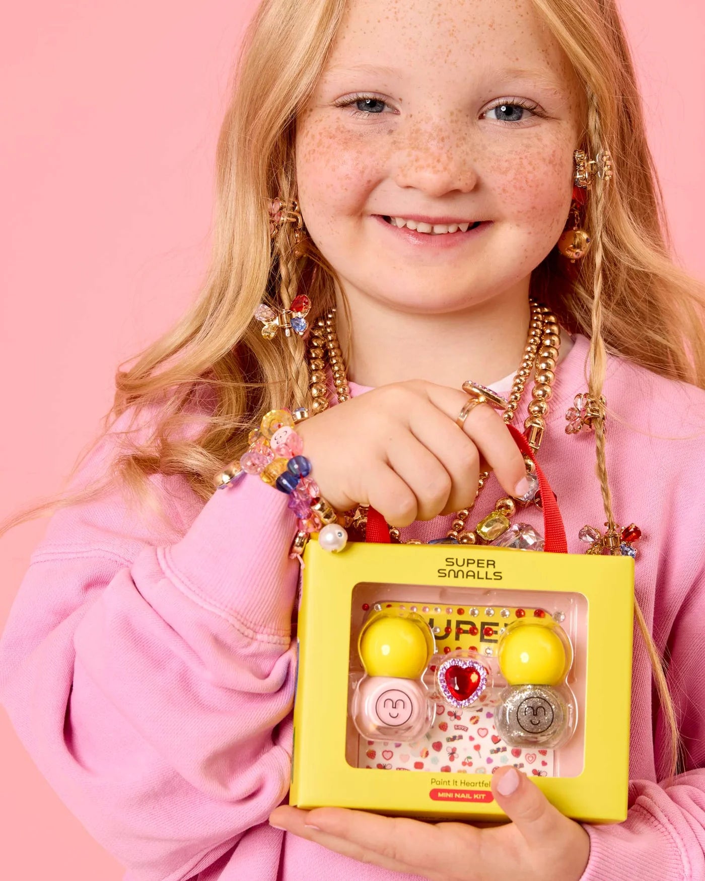 A smiling child with long blonde hair and colorful jewelry holds a yellow Super Smalls gift box containing lip gloss, rings, and the Paint it Heartfelt Mini Nail Kit against a pink background.