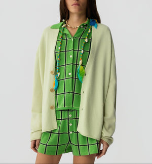 A person wearing a green checkered shirt and shorts set, layered with the Everywear Cardigan by Guest in Residence in light yellow, accessorized with a beaded necklace and feather charms.